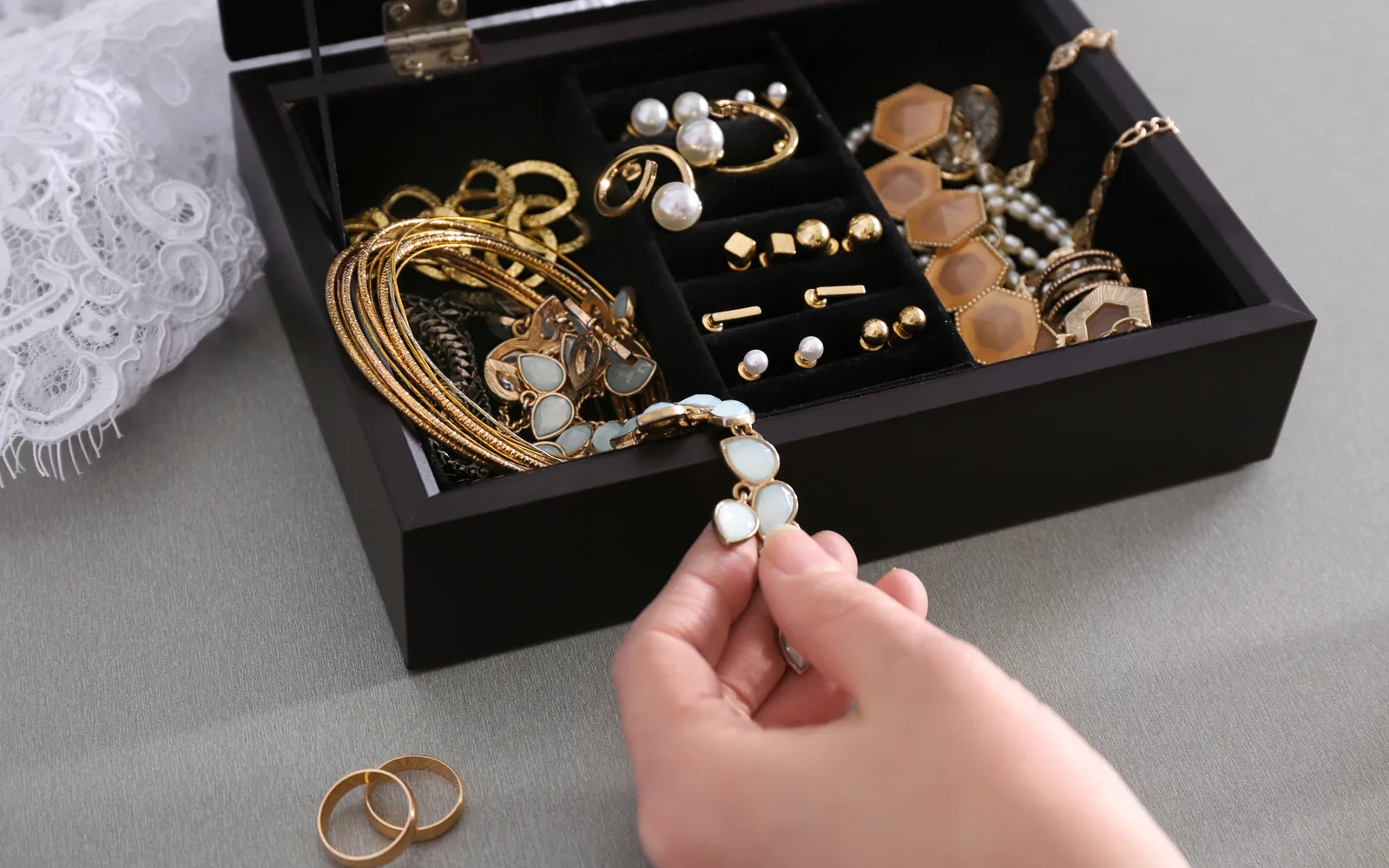 A jewelry box of gold bangles and pearl earrings, two gold bands beside it, one bracelet lifted out by hand