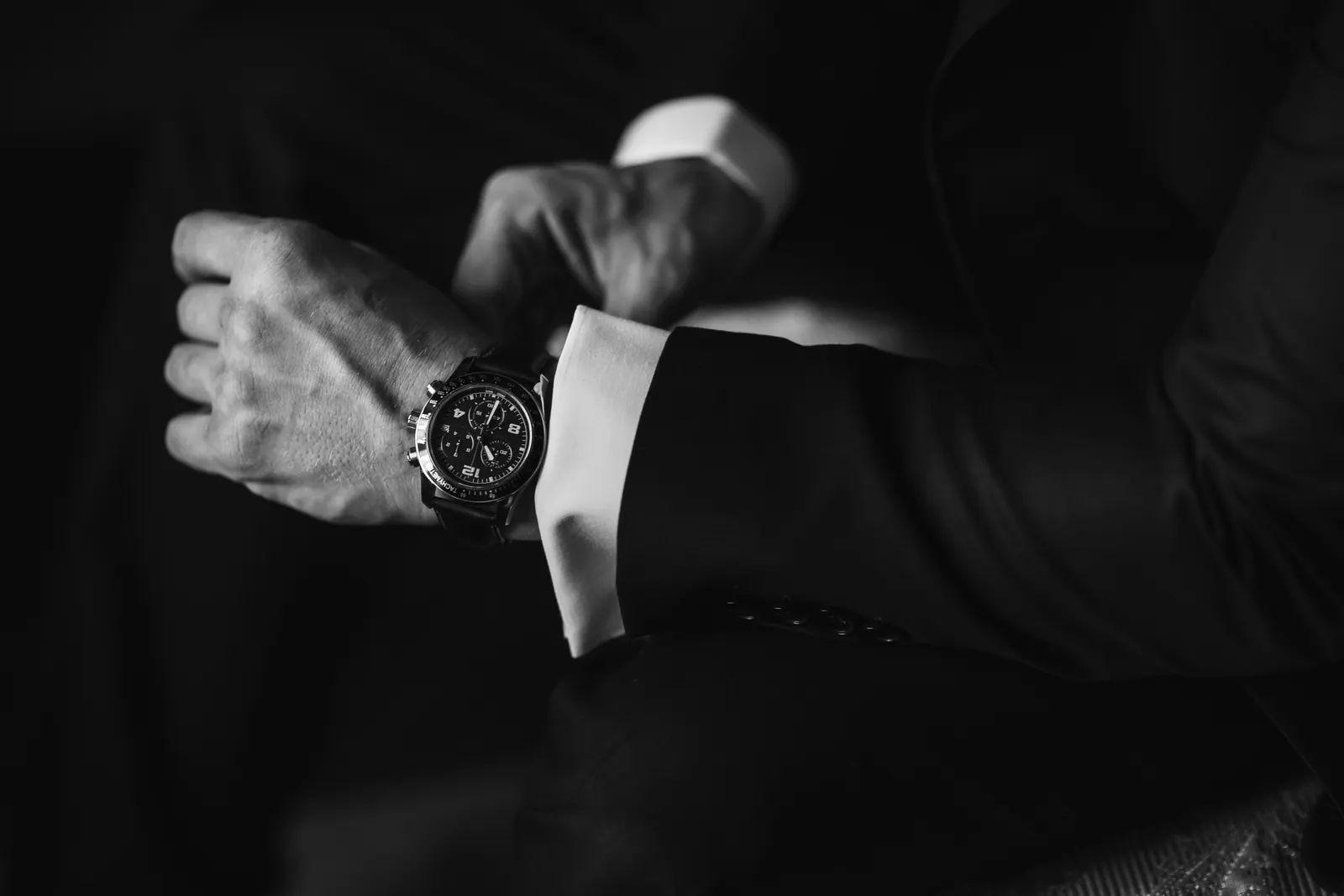 A man in a dark suit adjusting his cuff, a chronograph on a leather strap at his wrist